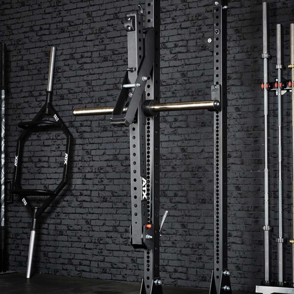 The gym setup showcases a black power rack and various barbells, including a trap bar and standard Olympic bars, against a dark brick wall. With the addition of the ATX® Belt Squat Option, it creates an organized and functional workout space.