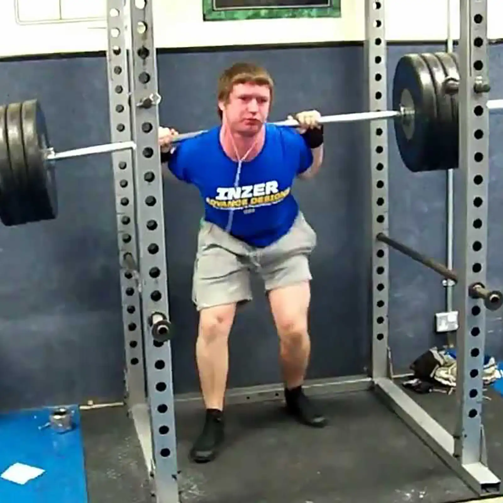A person wearing a blue shirt and gray shorts is squatting with a heavy barbell in a gym, near an ATX Compact Leg Press. They appear focused, using the power rack for support amid the blue walls and various equipment.