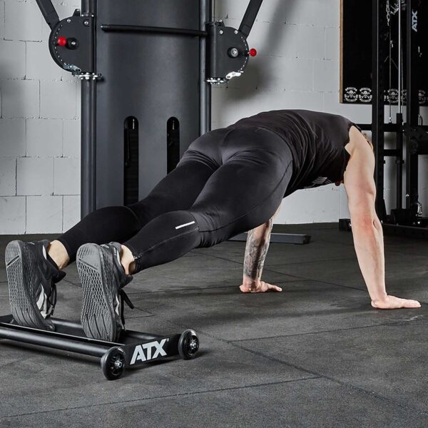 A person in a gym performs a decline push-up using a low bench, wearing black athletic clothing and shoes. The gym features various equipment, including the ATX® Glute Ham Roller visible in the background, with gray walls and rubber flooring.