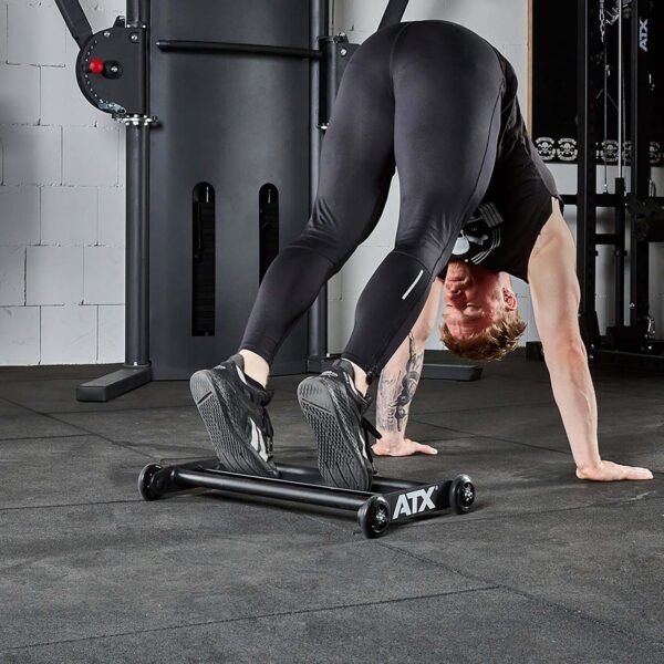 A person is performing a fitness exercise with the ATX® Glute Ham Roller in the gym. They are bent over with hands and feet on the floor, clad in black athletic clothing and shoes. Gym equipment, including a GHR machine, can be seen in the background.