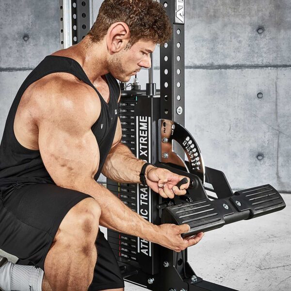 A man at a gym adjusts the ATX® Trinity Tower 135 kg. He wears a black tank top and shorts, concentrating on the machine. A concrete wall and a weights rack labeled Xtreme are visible in the background.