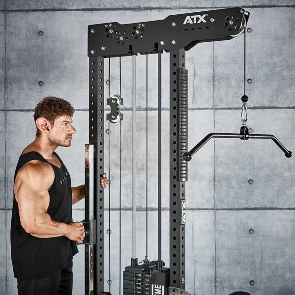 Wearing a black tank top, a person uses the ATX® Trinity Tower 135 kg, a tall black fitness machine with weights and pulley cables, against a concrete wall featuring a grid pattern.