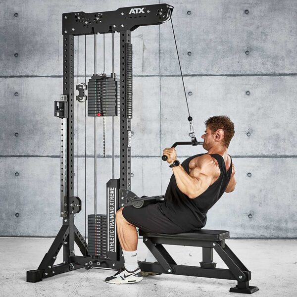 A person is using the ATX® Trinity Tower 135 kg for a seated lat pulldown in the gym. They wear a black tank top, black shorts, and white sneakers against a backdrop of a gray concrete wall.