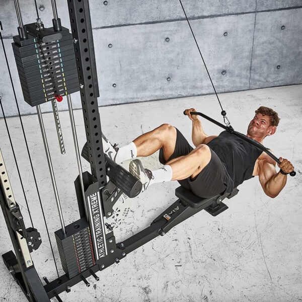 In a concrete room, a person on the ATX® Trinity Tower 135 kg is performing a rowing exercise by pulling a cable bar to their chest, feet secured on the platform. They are dressed in a black tank top, black shorts, and white socks.