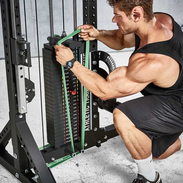 A muscular person in a black tank top and shorts adjusts a green resistance band on an ATX® Trinity Tower 135 kg weight stack machine in the gym, its setting confirmed by the equipment and concrete floor.