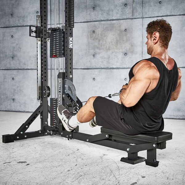 Someone with a muscular build is using the ATX® Trinity Tower 135 kg seated rowing machine in a gym. They wear a black tank top, black shorts, and white socks with sneakers against a backdrop of a concrete wall.