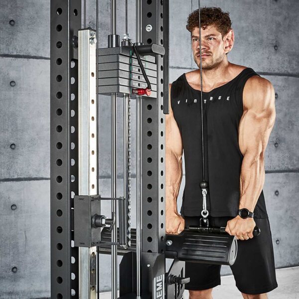 A person in a black tank top exercises on the ATX® Trinity Tower 135 kg, pulling down a bar with both hands. They are in a modern gym with a gray concrete wall backdrop.