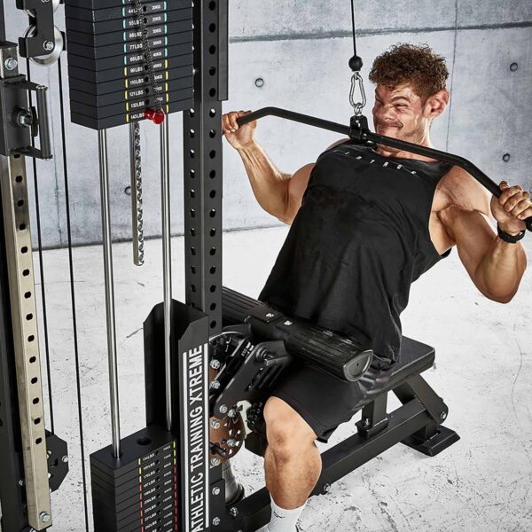 A person in a gym does a sitting lat pulldown on the ATX® Trinity Tower 135 kg cable machine, wearing a black tank top and shorts. The gym equipment and weights are prominently visible.