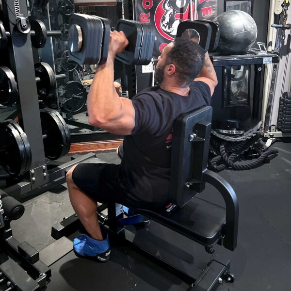 Seated on an Ironmaster Seated Press Pad SB in a gym, a person lifts dumbbells overhead, dressed in a black shirt and shorts with blue shoes. Various gym equipment like weights and a medicine ball are visible in the background.