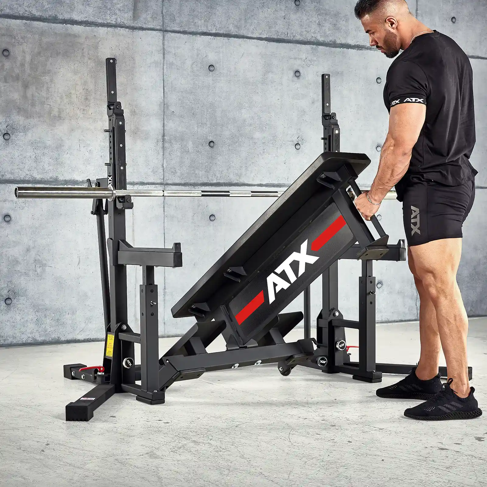 A muscular man in black athletic wear adjusts an adjustable gym bench with a barbell in a concrete-walled gym. The bench, part of an IPF combo rack branded ATX, is essential for his competition bench press setup, and he is focused on setting up the equipment.