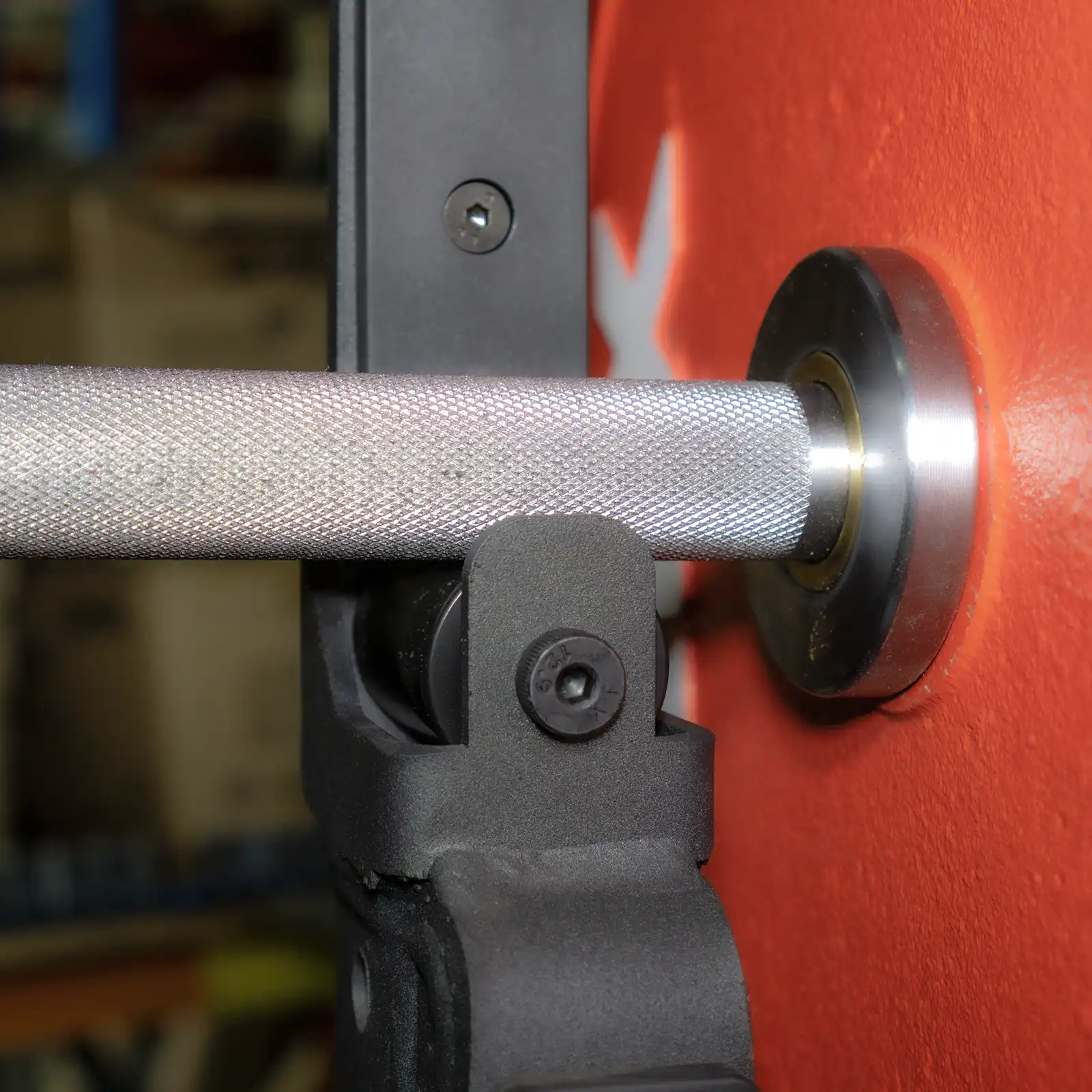 Close-up of a barbell resting on an IPF combo rack against an orange wall. The metal bar features a textured grip, and the sturdy black and silver components enhance its robust design. The background is slightly blurred, capturing the essence of an energetic gym setting.