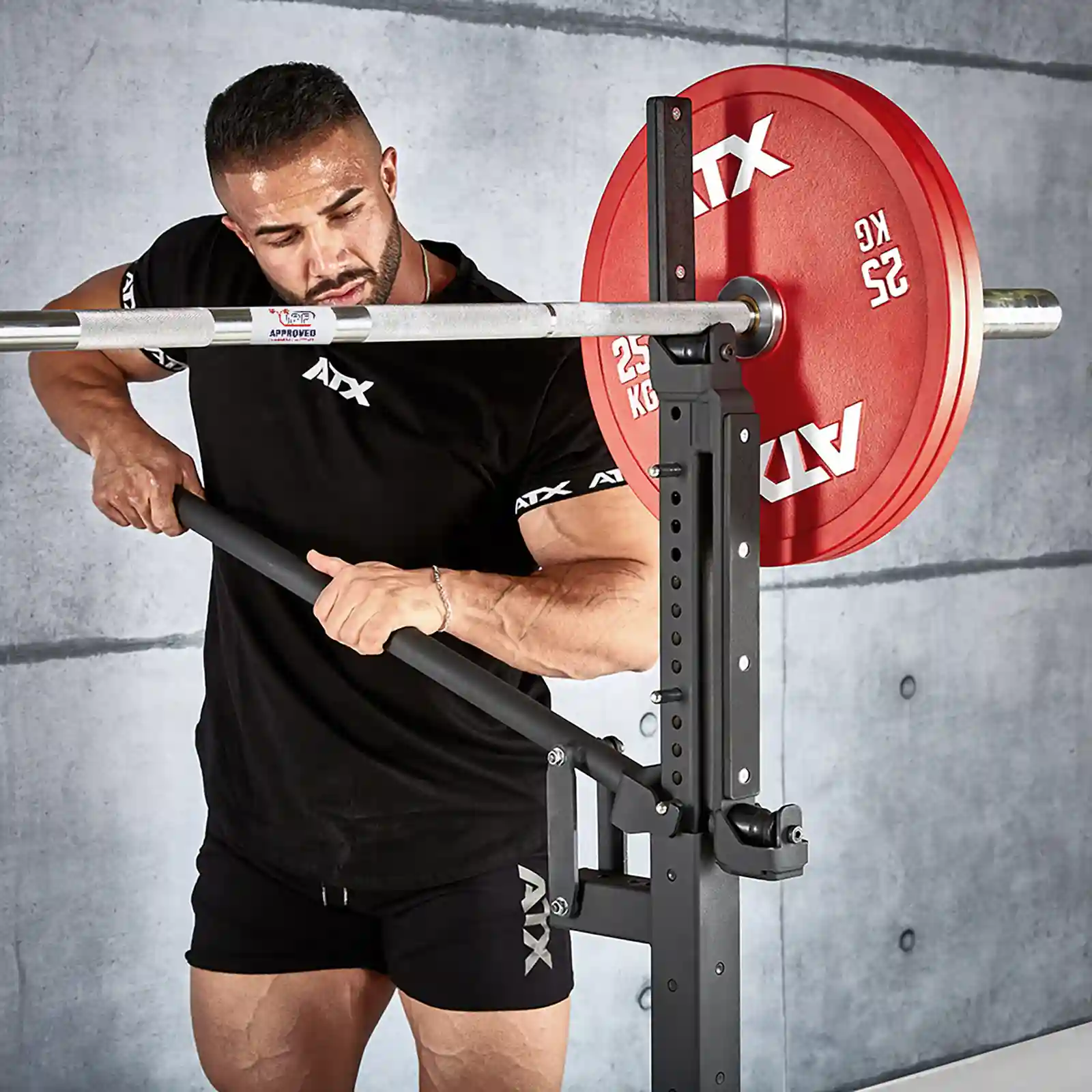 A man wearing a black ATX t-shirt and shorts is adjusting an IPF combo rack with a red weight plate, seamlessly blending squat rack and bench elements. The background features a gray concrete wall.