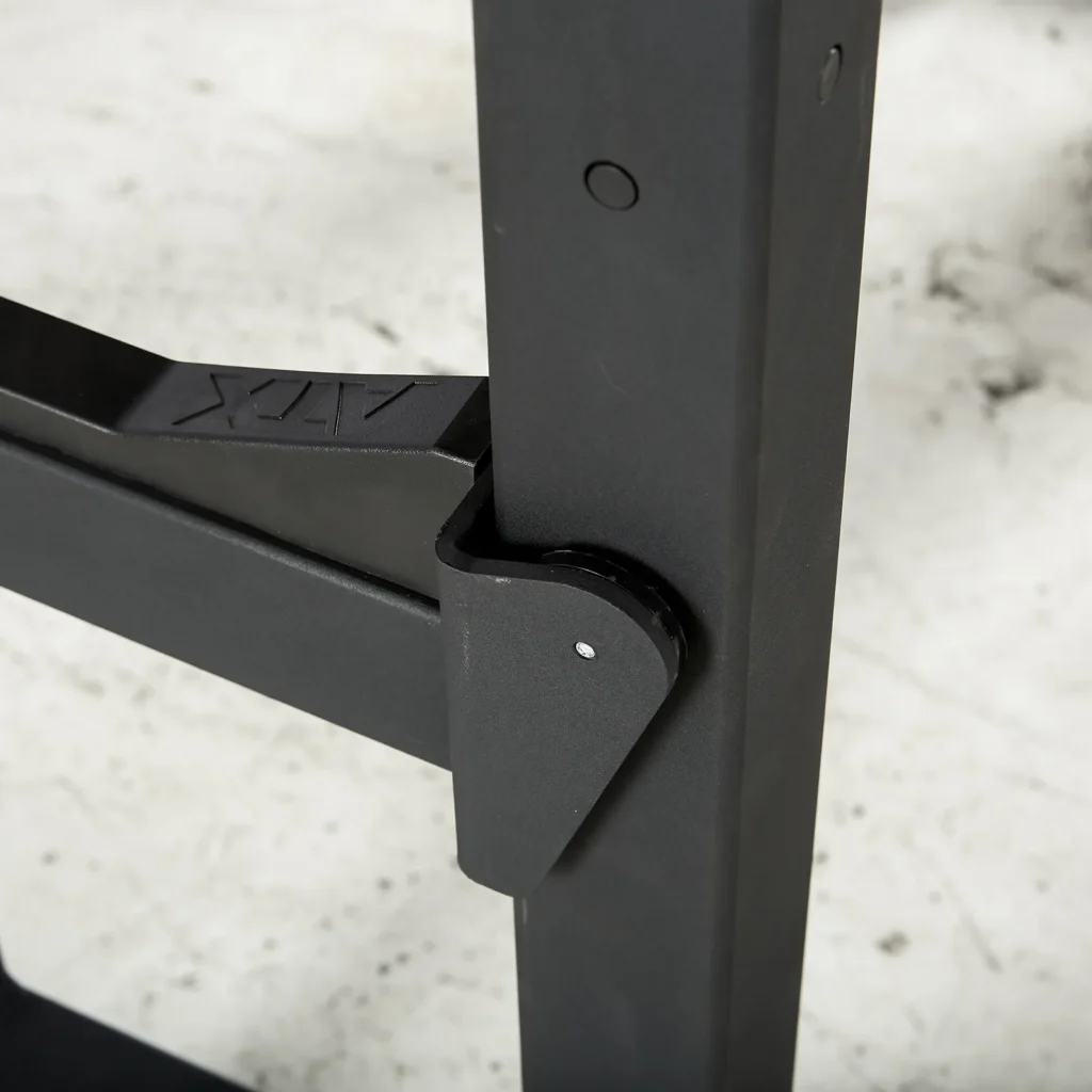 Close-up of a black metal hinge on equipment, resting on a concrete floor. Connected to a rectangular structure with a visible bolt and subtle engraved logo, this component is reminiscent of those found in an IPF combo rack, bridging design with strength.