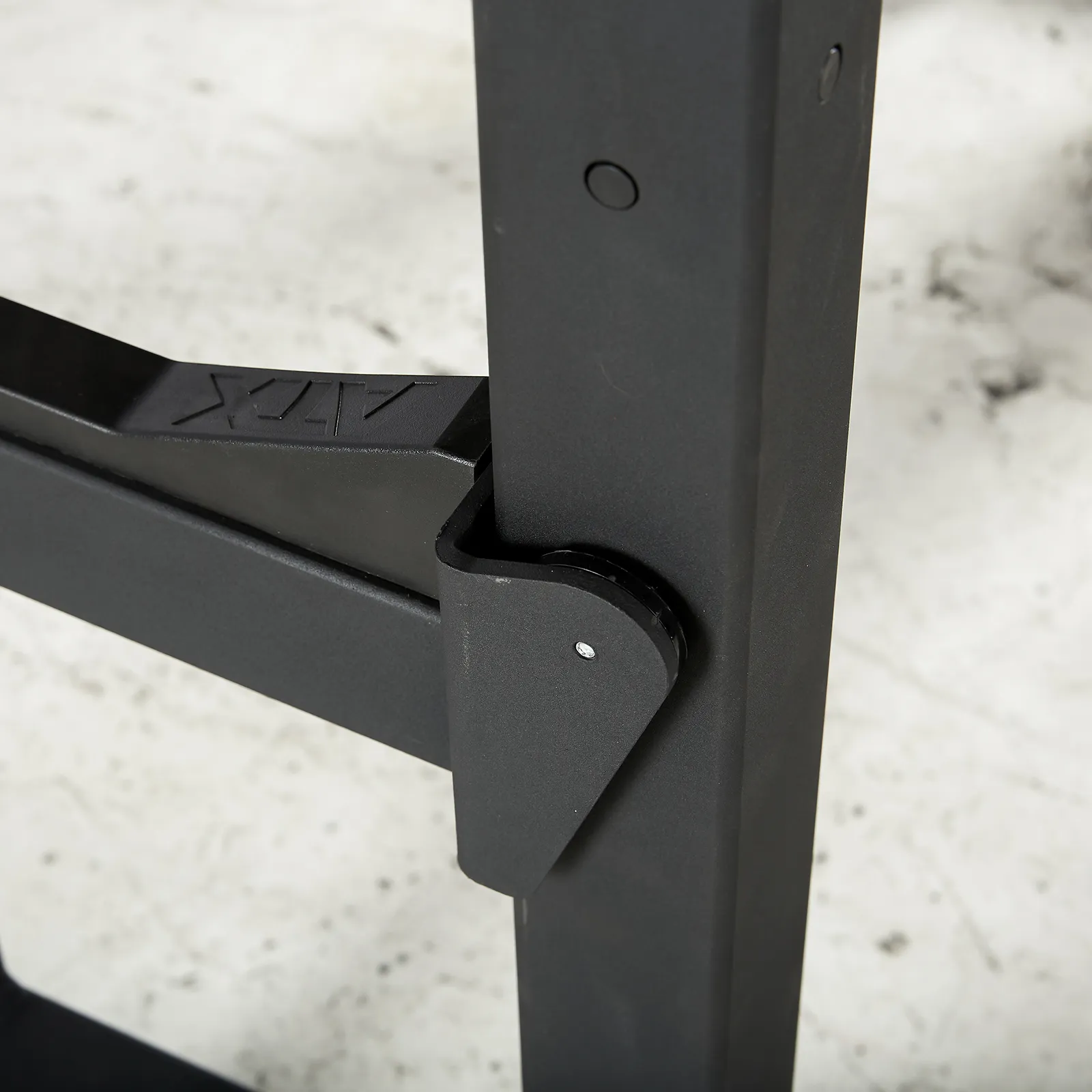 Close-up of a black metal hinge on equipment, resting on a concrete floor. Connected to a rectangular structure with a visible bolt and subtle engraved logo, this component is reminiscent of those found in an IPF combo rack, bridging design with strength.