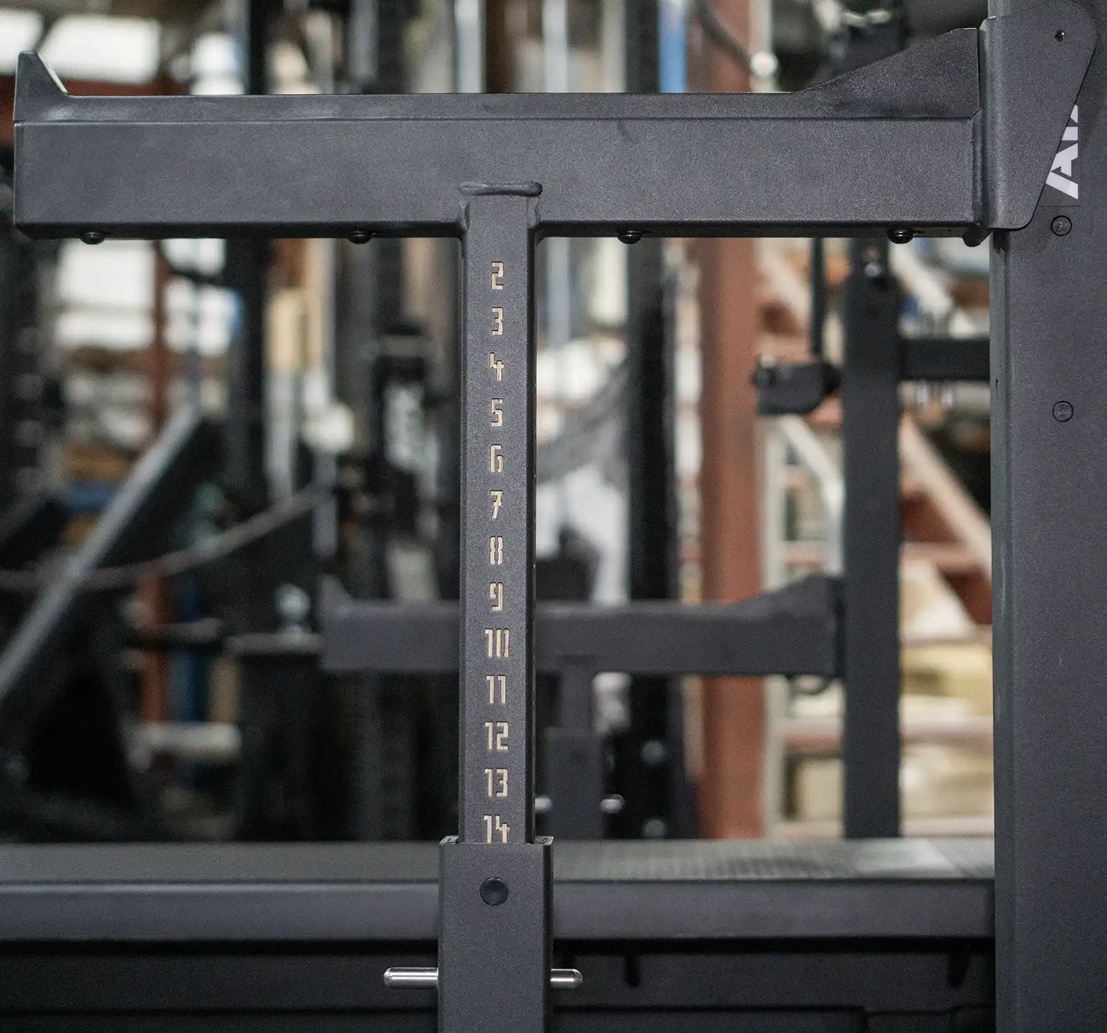 Close-up of a squat rack and bench with numbered height levels from 2 to 13 on a vertical bar. The background is blurred, showcasing gym equipment and a staircase, creating the perfect setting for serious lifters or those prepping for competition.