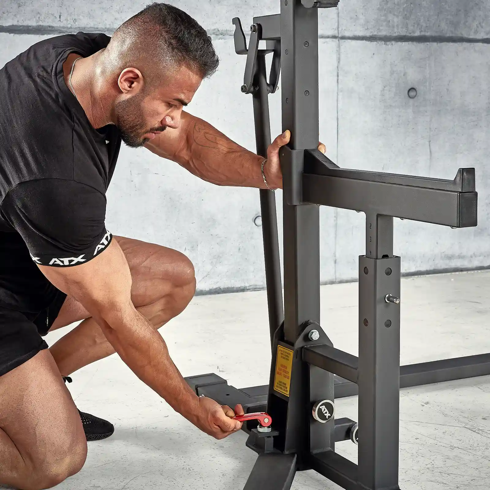 A man adjusts a lever on an IPF combo rack in a modern indoor gym with concrete walls. Focused on modifying the settings, he wears a black workout outfit and shoes, ensuring optimal performance during his session.