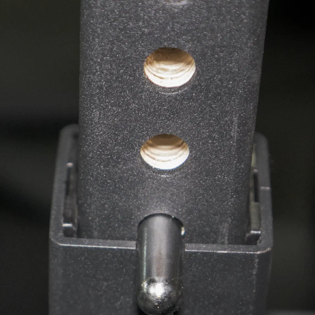 Close-up of a metal frame with two circular holes and a protruding pin, showcasing the matte, dark texture typical of an IPF combo rack. Likely part of an adjustable mechanism or piece of equipment designed for versatile lifting setups.