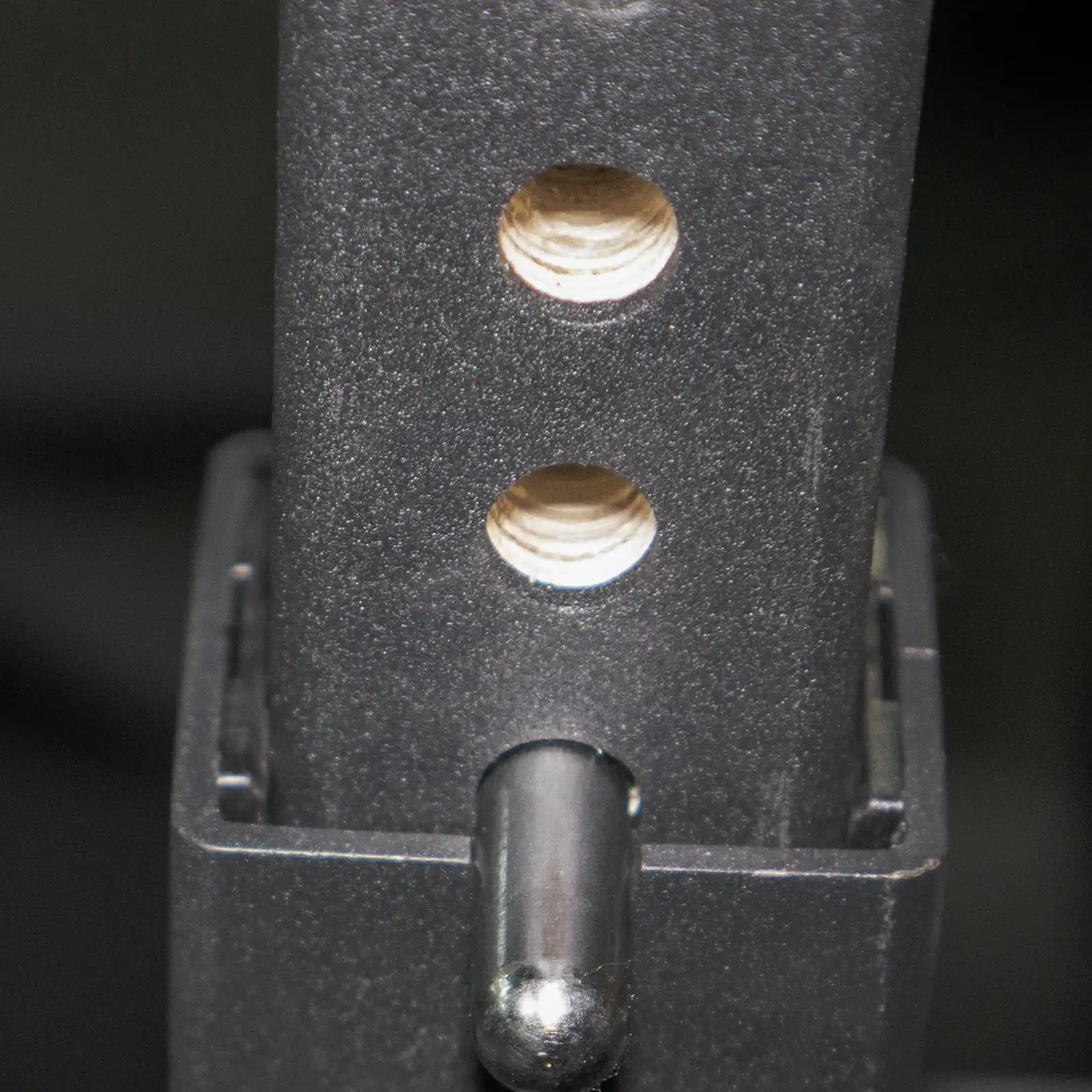 Close-up of a metal frame with two circular holes and a protruding pin, showcasing the matte, dark texture typical of an IPF combo rack. Likely part of an adjustable mechanism or piece of equipment designed for versatile lifting setups.