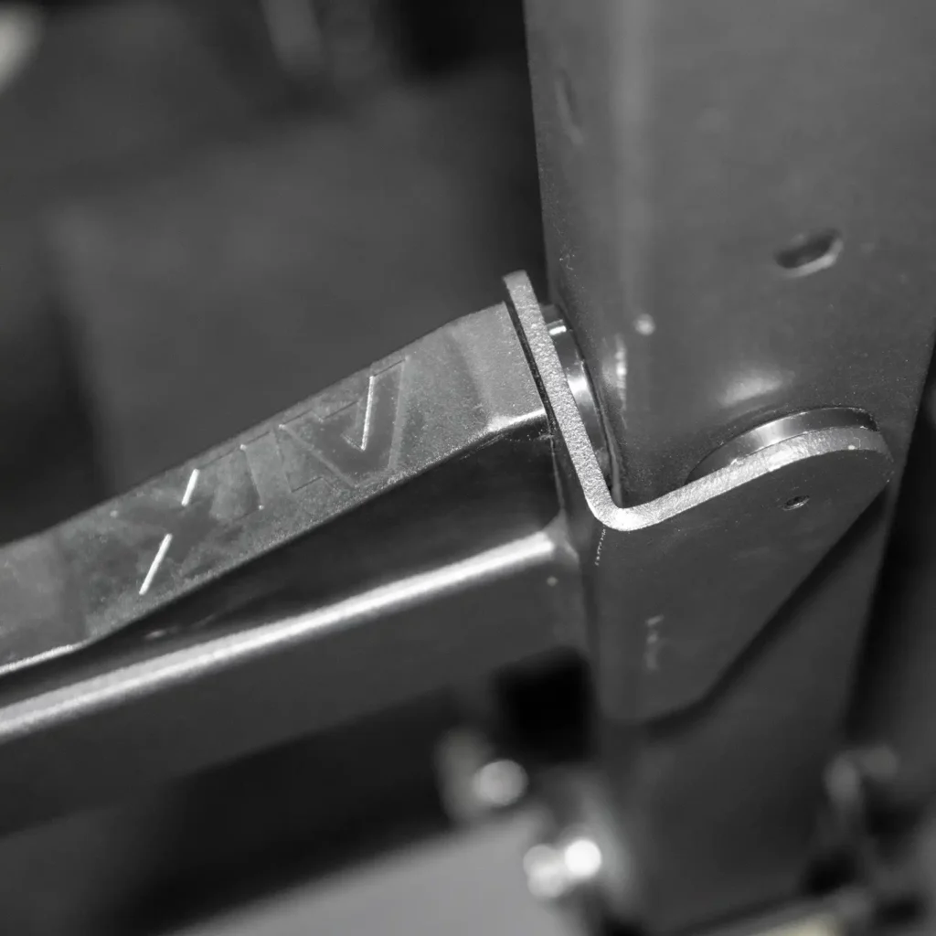 Close-up of a metal hinge joint connecting parts of a squat rack, with a bolt securing it. Made of sturdy, dark metal, it features an engraved AV for branding. This design ensures reliability whether youre using the squat rack and bench for intense workouts or in competition settings.