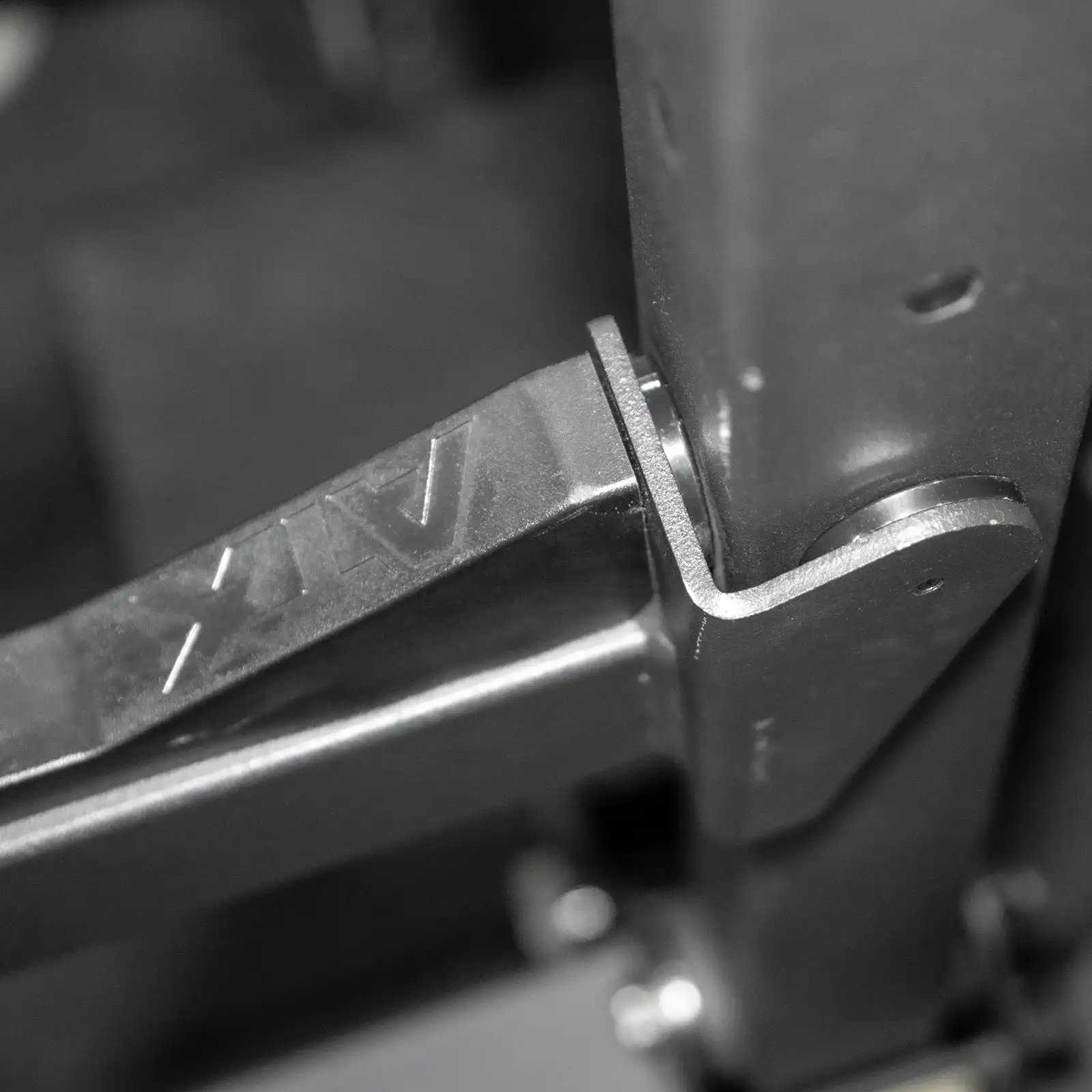 Close-up of a metal hinge joint connecting parts of a squat rack, with a bolt securing it. Made of sturdy, dark metal, it features an engraved AV for branding. This design ensures reliability whether youre using the squat rack and bench for intense workouts or in competition settings.