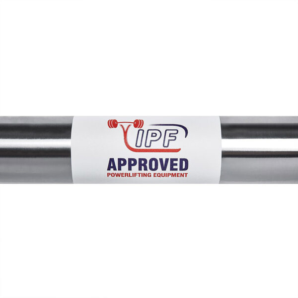 A close-up of a metallic bar with a white label displaying the IPF logo, featuring a red barbell graphic and the text APPROVED POWERLIFTING EQUIPMENT in bold red and blue letters on the ATX® Warrior Power Bar IPF.