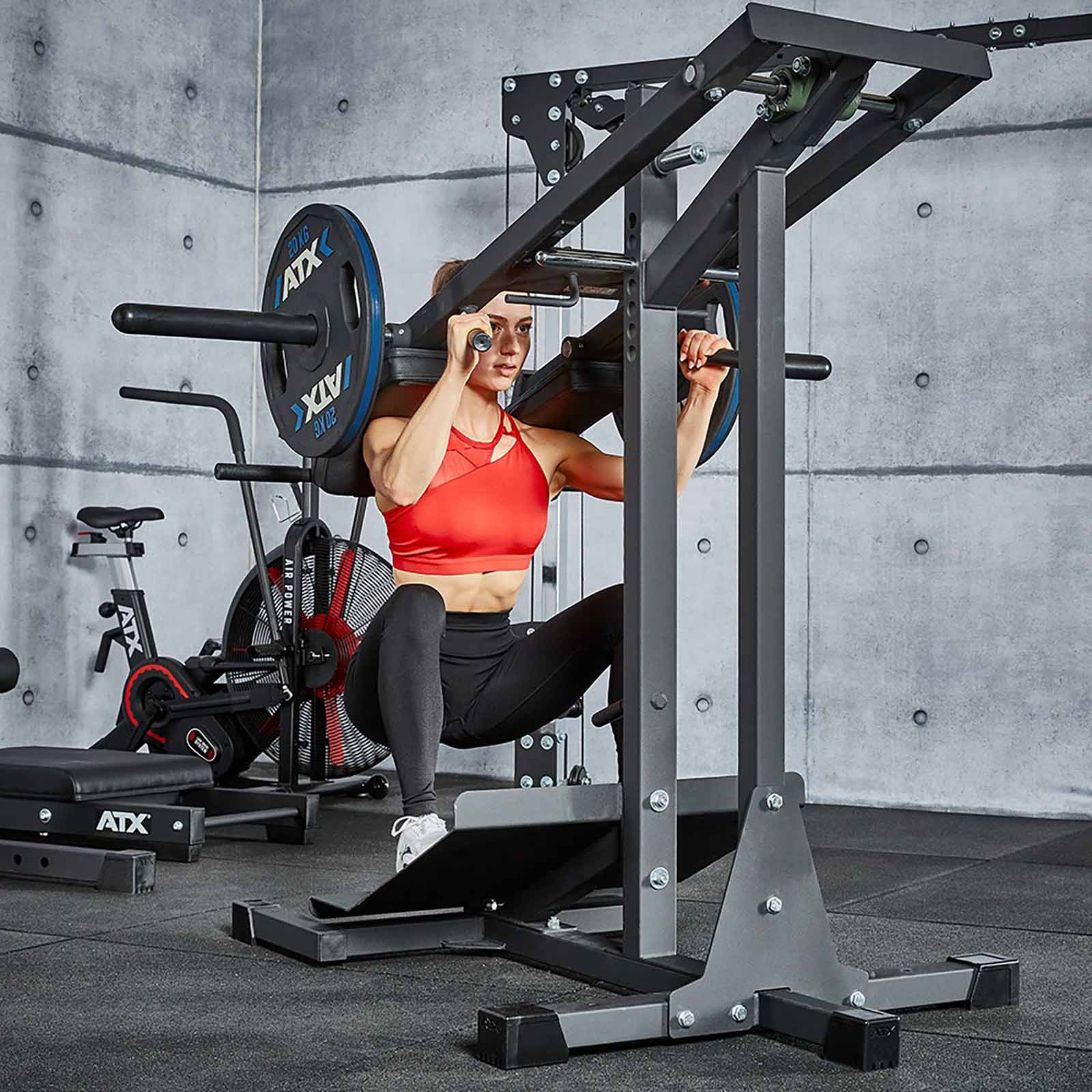 A person in a red sports bra and black leggings expertly performs a squat on the leverage squat machine at the gym. Behind them, weights and various exercise equipment add to the focused workout atmosphere.