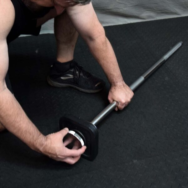 A person in a black tank top and sneakers kneels on a black mat, placing a weight plate onto the Ironmaster Quick Lock Mace Handle.