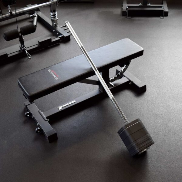 An Ironmaster Quick Lock Mace Handle is placed diagonally across a flat black weight bench on a gym floor, with other gym equipment visible in the background.