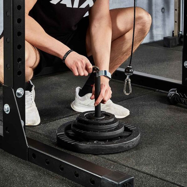 Next to the Cable Pulley System (High), a person in a black tank top, shorts, white sneakers, and a wristwatch adjusts weights on the loading pin. The gyms black mats cover the floor.