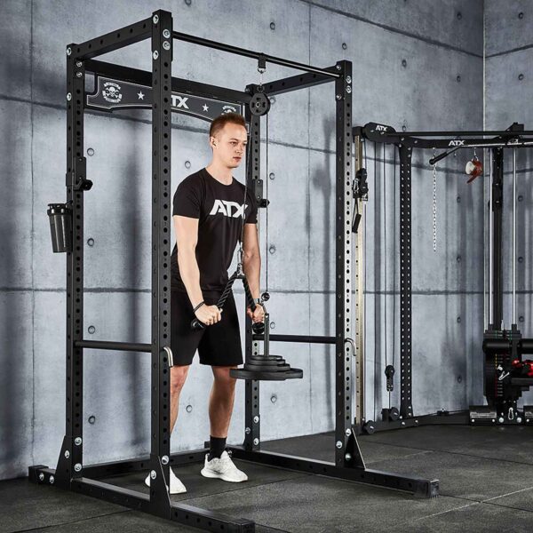 A person, in a black t-shirt and shorts, exercises using the Cable Pulley System (High) inside a metal frame at the gym. The concrete-walled gym also features other workout equipment in the background.