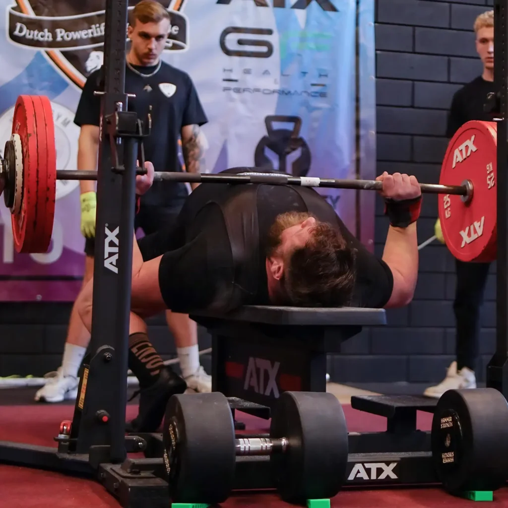 At a powerlifting event, a person executes a competition bench press on an IPF combo rack, lifting a heavily weighted barbell. Two spotters stand ready for safety as various banners and gym equipment form the backdrop.