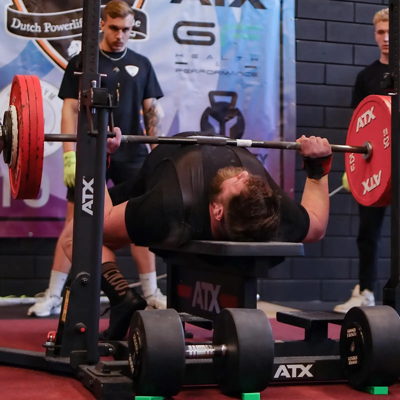 At a powerlifting event, a person executes a competition bench press on an IPF combo rack, lifting a heavily weighted barbell. Two spotters stand ready for safety as various banners and gym equipment form the backdrop.
