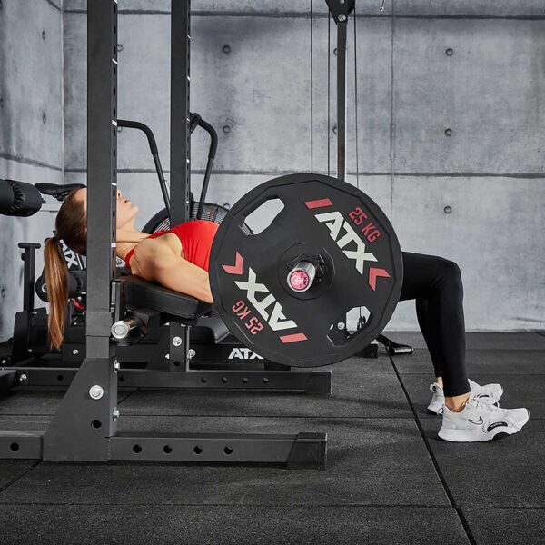 A long-haired person in a red top and black leggings skillfully performs a hip thrust exercise with a weighted barbell using the ATX® Hip Thruster Attachment. The gym features concrete walls and black flooring, and the weights are marked ATX 25 KG.