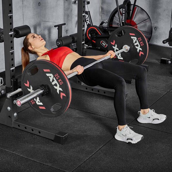 A person in a red sports bra and black leggings uses the ATX® Hip Thruster Attachment to perform a hip thrust exercise with perfect form, positioning themselves on a bench with feet flat while the barbell rests securely on the platform.