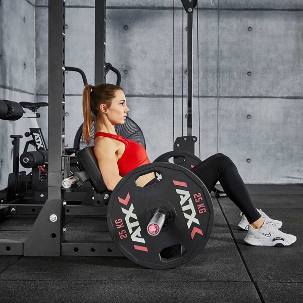 A woman in a red top and black leggings performs a leg press with the ATX® Hip Thruster Attachment at the gym. She presses 25 kg on the platform, surrounded by modern, industrial gym equipment.