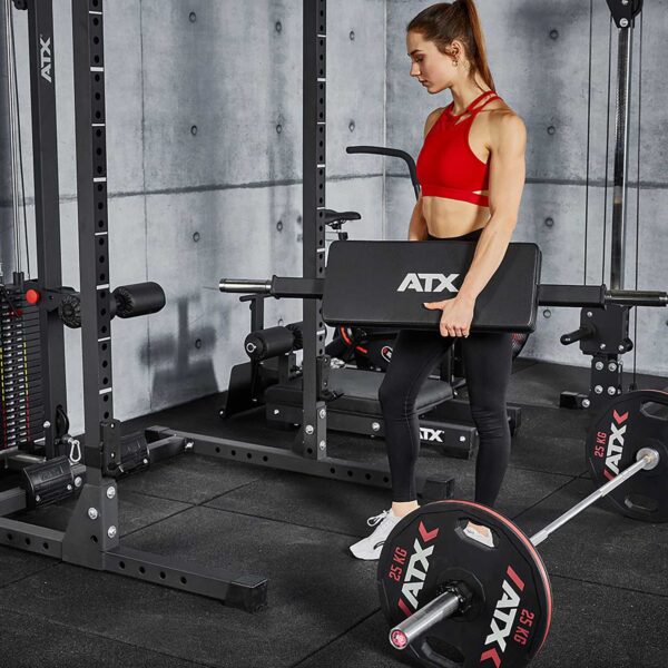 In a gym with a concrete wall, a person in a red sports bra and black leggings holds an ATX padded board, with the ATX® Hip Thruster Attachment and a loaded barbell nearby among other fitness equipment.