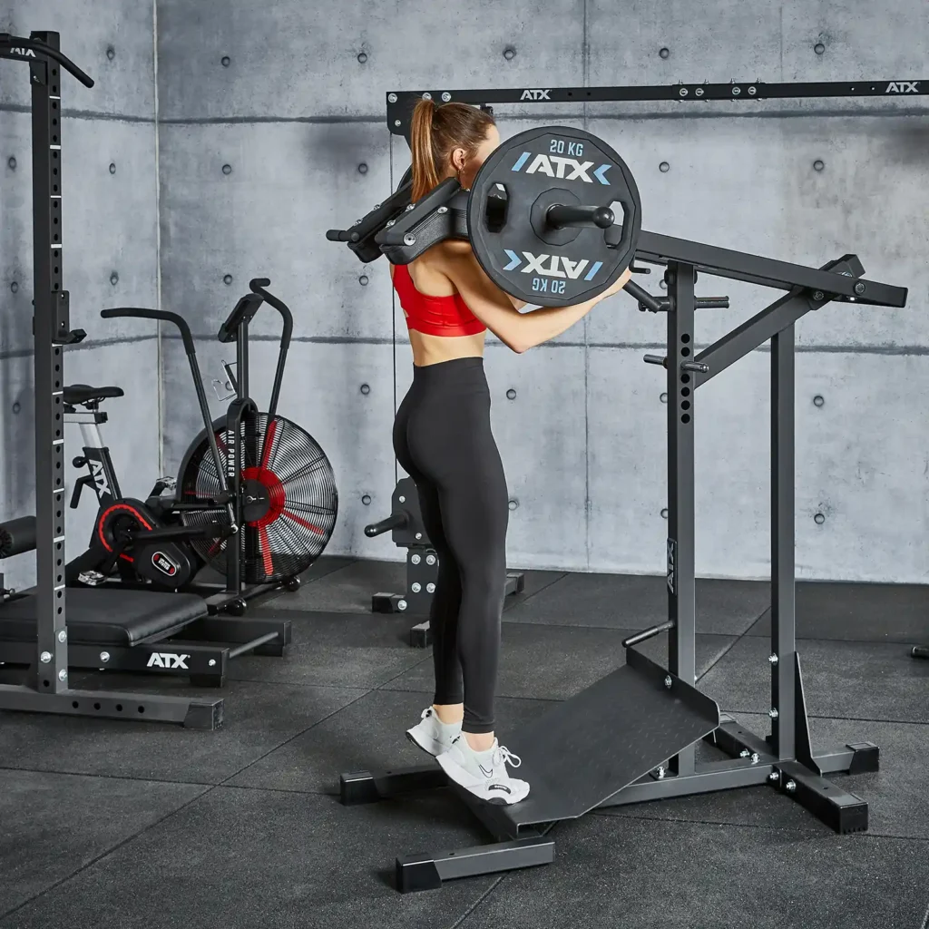 A woman in a red sports bra and black leggings uses a calf raise machine in a gym, where fitness equipment like a leverage squat machine, fan bike, and squat rack dot the space. The rooms concrete walls add an industrial edge to this fitness haven.