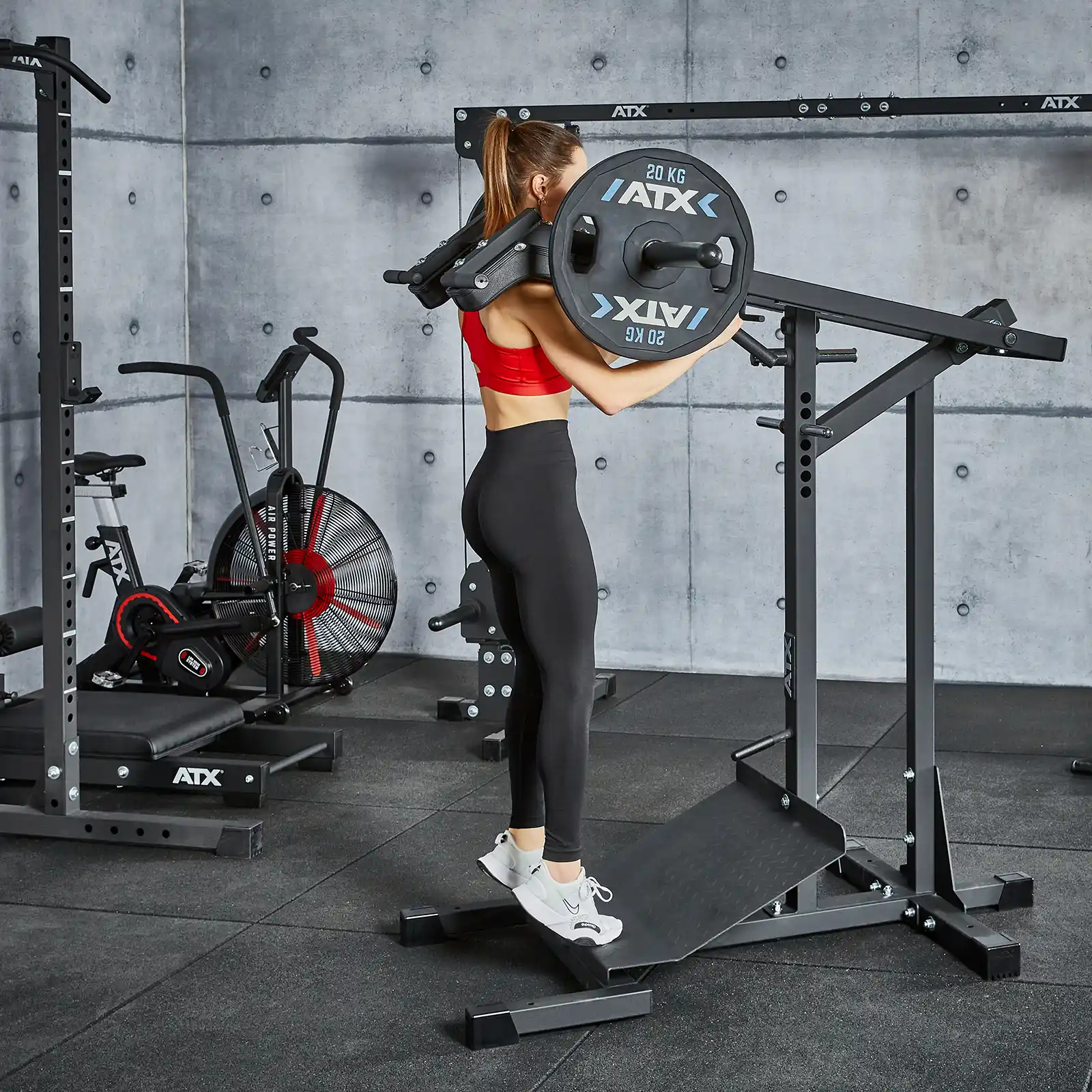 A woman in a red sports bra and black leggings uses a calf raise machine in a gym, where fitness equipment like a leverage squat machine, fan bike, and squat rack dot the space. The rooms concrete walls add an industrial edge to this fitness haven.