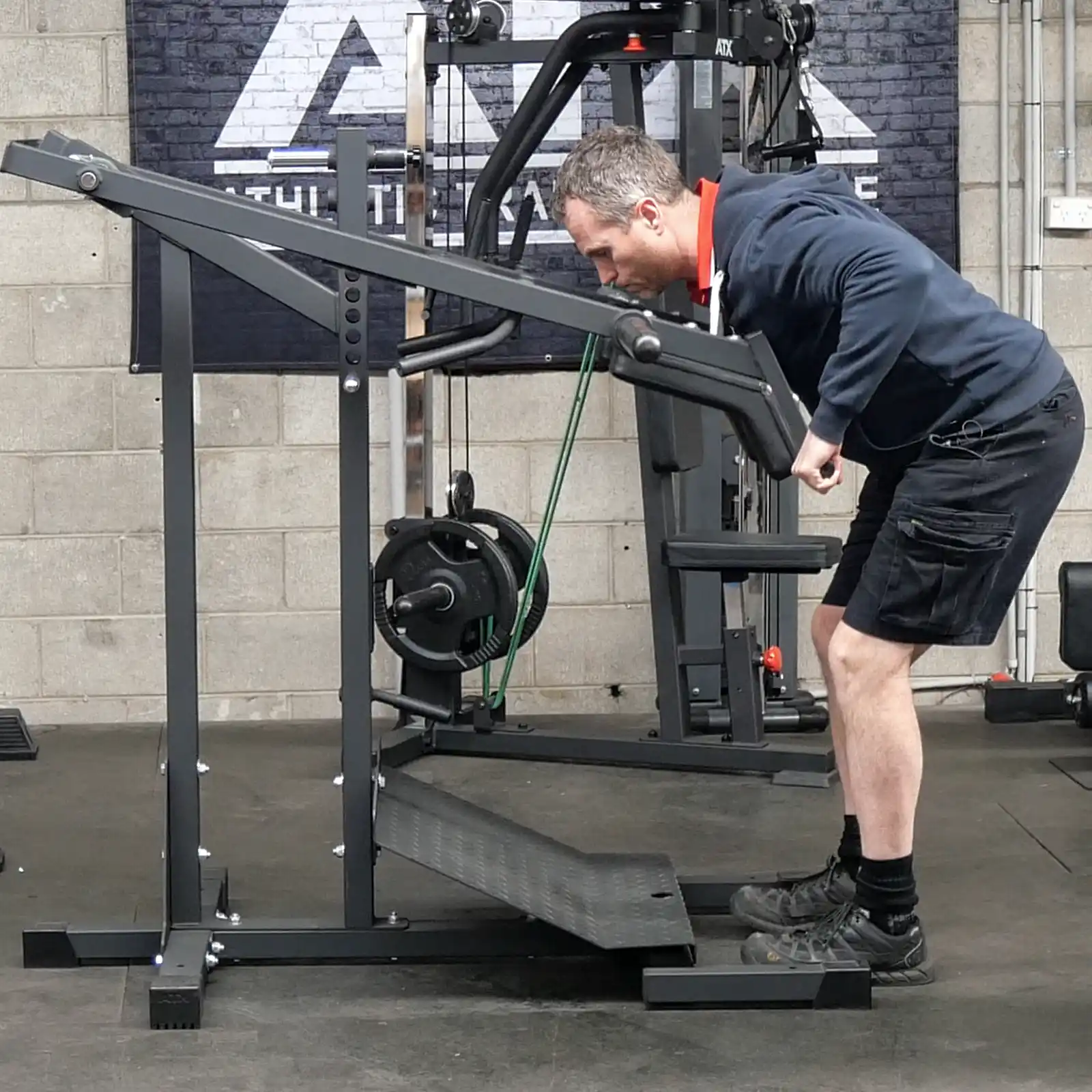 A man in a gym is using a leverage squat machine for exercising. He is bending forward slightly while holding onto handles attached to weights. The background features gym equipment and a brick wall with a banner.