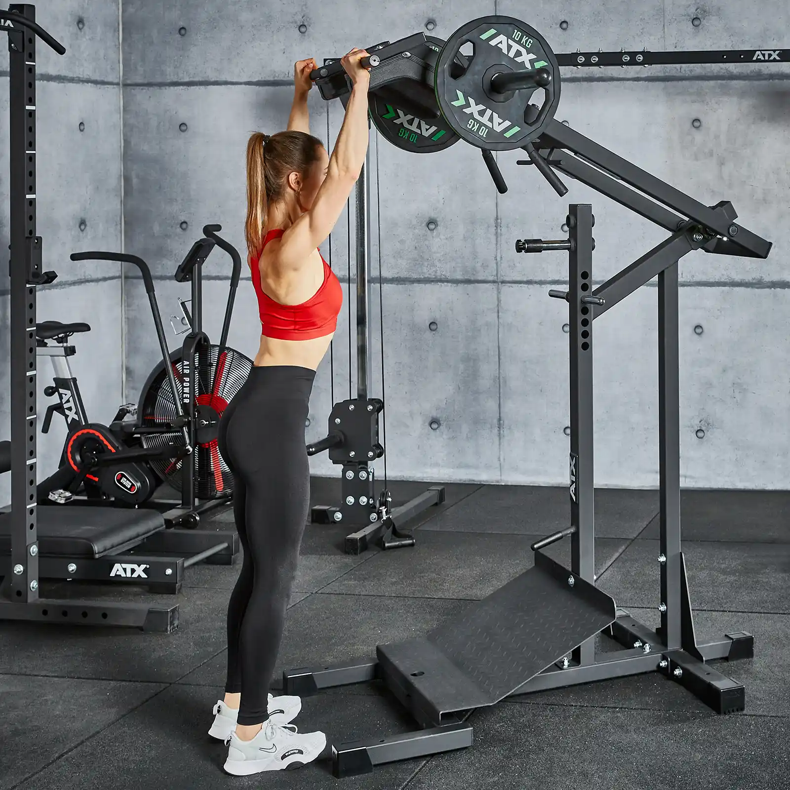 A person in athletic clothing performs a shoulder press using a weightlifting machine in a modern gym, where gray concrete walls and a leverage squat machine populate the backdrop amidst various exercise equipment.
