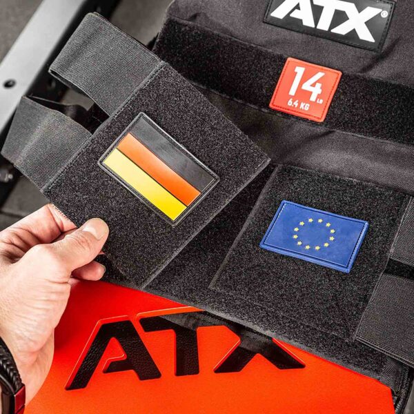A hand holds a German flag patch on the ATX® Tactical Weight Vest, featuring another patch with the EU flag. A 14 kg weight label is visible. The vest rests on a red and black surface with ATX branding.