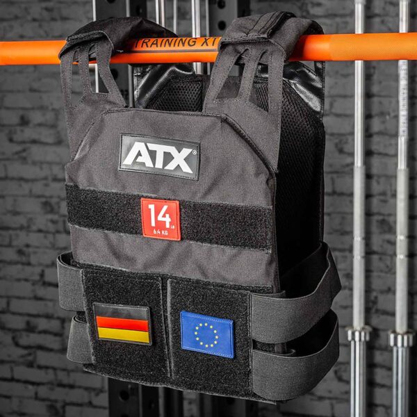 An ATX® Tactical Weight Vest with a 14 kg patch and flags of Germany and the EU hangs on an orange bar against a gray brick wall with metal rods.