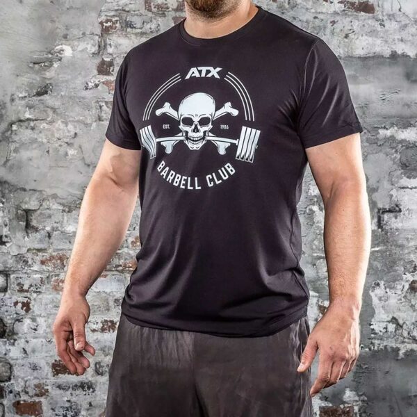 A person in black ATX® Barbell Club T-Shirt, featuring a skull and crossbones, stands in front of a weathered brick wall, paired with black shorts.