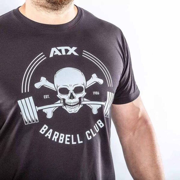 A person models the ATX® Barbell Club T-Shirt Black, showcasing a skull with crossed bones and a barbell in its mouth. The prints say ATX Barbell Club and Est. 1986. The shirt is displayed against a white background.