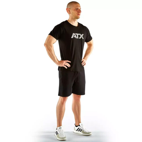 A person stands confidently with hands on hips, wearing a black ATX® Training Shirt, black shorts, and white Adidas sneakers against a plain white background.