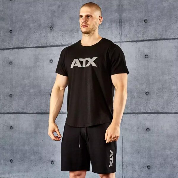 A man stands in a gym wearing the ATX® Training Shirt BLACK and matching shorts with ATX printed on them. The background features a gray concrete wall with a modern industrial design, and he appears ready for a workout.