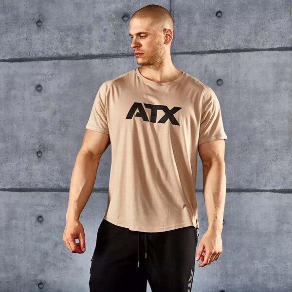 A man in a beige T-shirt with ATX printed on it stands against a concrete wall. He is looking to the side and wearing black athletic pants.
