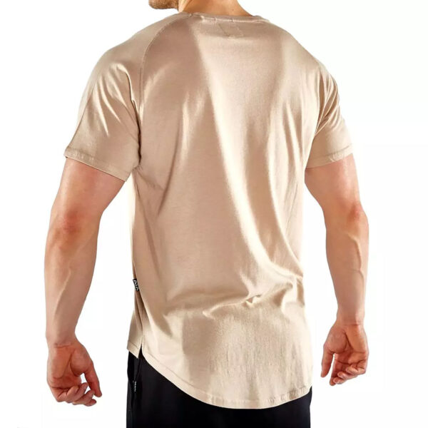 A person wearing a beige short-sleeve t-shirt and black pants, viewed from the back, stands against a plain white background. The t-shirt has a curved hem. The person has short hair and their hands are relaxed by their sides.