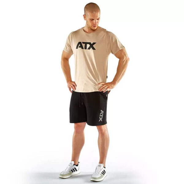 A man stands wearing a beige t-shirt with ATX printed on it, black shorts with a small ATX logo, and white sneakers with black stripes. He is looking down with hands on hips against a plain white background.