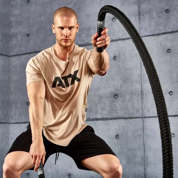 A muscular man exercises with battle ropes in a gym setting. He wears a beige ATX t-shirt and black shorts, with a focused expression. The concrete wall background adds to the rugged, intense workout atmosphere.